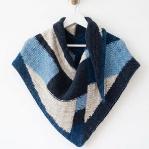 May include: A triangular knit shawl featuring a geometric colour-blocked pattern in shades of navy blue, light blue, and cream. The accessory is constructed with a textured garter stitch and is displayed hanging from a white plastic clothes hanger.