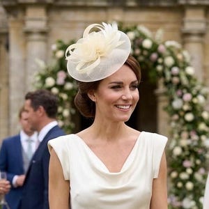 Könnte beinhalten: Ein cremefarbenes, bodenlanges Kleid mit Wasserfall-Ausschnitt und kurzen Ärmeln, kombiniert mit einem passenden cremefarbenen Fascinator mit zarten Federakzenten und einer kleinen, strukturierten cremefarbenen Clutch in der Hand.