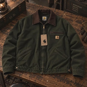 May include: A dark olive green canvas jacket featuring a brown corduroy collar, a full-length front zipper, and two slanted lower pockets. A Carhartt brand tag hangs from the zipper pull, and a small logo patch is on the left chest pocket.