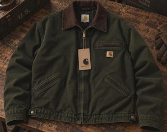 May include: A dark olive green canvas jacket featuring a brown corduroy collar, a full-length front zipper, and two slanted lower pockets. A Carhartt brand tag hangs from the zipper pull, and a small logo patch is on the left chest pocket.