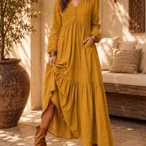 May include: A mustard yellow long-sleeved maxi dress featuring a V-neckline with a vertical row of small buttons, a gathered waistline, side pockets, and a tiered skirt with a ruffled hemline, paired with brown leather ankle boots.