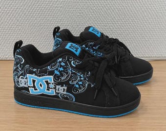 May include: Black DC trainers featuring a light blue and white swirling graphic pattern on the side panels. The shoes have black laces, a padded tongue with a blue DC logo patch, and a solid black midsole with a bright blue rubber outsole.