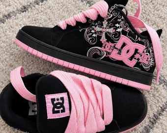 May include: A pair of black and pink DC sneakers featuring thick pink laces, a pink DC logo on the tongue and side, and a silver cross charm attached to the eyelet. The side panel displays a white floral pattern, and the sole is pink.