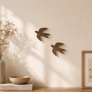 Puede incluir: Dos siluetas de golondrinas de madera de color marrón oscuro montadas en una pared. Las aves se representan en vuelo con las alas extendidas, con superficies talladas lisas y patrones de vetas de madera natural. Estas decoraciones de pared están colocadas en diagonal.
