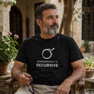 以下が含まれることがあります： 黒の半袖Tシャツ。胸元には、タッセルが付いた円形のビーズチェーンの白いグラフィックと、白い大文字で「REMEMBRANCE IS RECURSIVE」というテキストがプリントされています。
