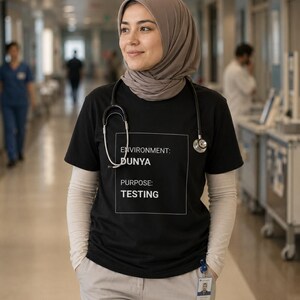 May include: A black short-sleeved t-shirt featuring a white square graphic with the text ENVIRONMENT: DUNYA and PURPOSE: TESTING. The shirt is worn over a long-sleeved beige base layer, paired with light-colored trousers and a stethoscope around the neck.