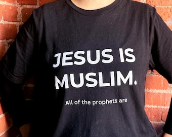 Può includere: Una t-shirt nera a maniche corte con testo bianco stampato sul davanti. Il testo recita "JESUS IS MUSLIM." in grandi lettere maiuscole, seguito dal testo più piccolo "All of the prophets are" posizionato direttamente sotto la frase principale.