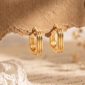 Puede incluir: Un par de pendientes de aro dorados con forma rectangular y tres crestas verticales distintas a lo largo de la superficie exterior. Los pendientes presentan un acabado metálico pulido y un mecanismo de cierre articulado.