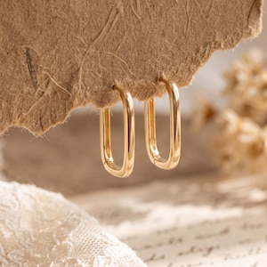 May include: A pair of gold-toned hoop earrings with a rectangular, elongated shape. The earrings feature a smooth, polished metallic finish and are suspended from a textured, light brown paper edge, showcasing their simple, open-loop design.