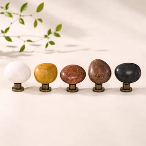May include: A set of five cabinet knobs featuring smooth, rounded, pebble-shaped tops in white, yellow, reddish-brown, brown, and black stone, each mounted on a short, cylindrical, metallic brass-toned base for attachment to furniture.