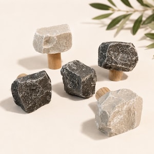 May include: A set of five cabinet knobs featuring raw, jagged stone tops in shades of light grey and dark charcoal, each securely attached to a cylindrical, light-coloured wooden base. The stones have irregular, natural textures and sharp edges.
