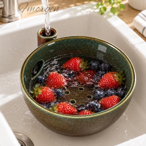 Könnte beinhalten: Eine runde, gesprenkelte dunkelgrüne Keramikschüssel als Beeren-Seiher, gefüllt mit frischen Erdbeeren und Blaubeeren, die unter einem Wasserhahn abgespült werden. Die Schüssel hat einen kleinen integrierten Griff und einen perforierten Boden, mit dem Wort "Fmorgen" in Schreibschrift an der Seite.