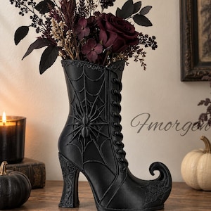 Könnte beinhalten: Eine mattschwarze stiefelförmige Vase mit einem erhabenen Spinnen- und Spinnennetzdesign an der Seite, Schnürdetails und einer geschwungenen Spitze. Die Vase ist mit einem Strauß dunkelroter Rosen und Trockenblumen gefüllt, Text Fmorgen sichtbar.