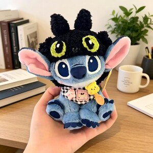 May include: A blue plush character toy wearing a black textured hat with green and black eye patterns. The toy features large pink-lined ears, a black and white checkered neck scarf with small star and bear charms, and a yellow spoon accessory.