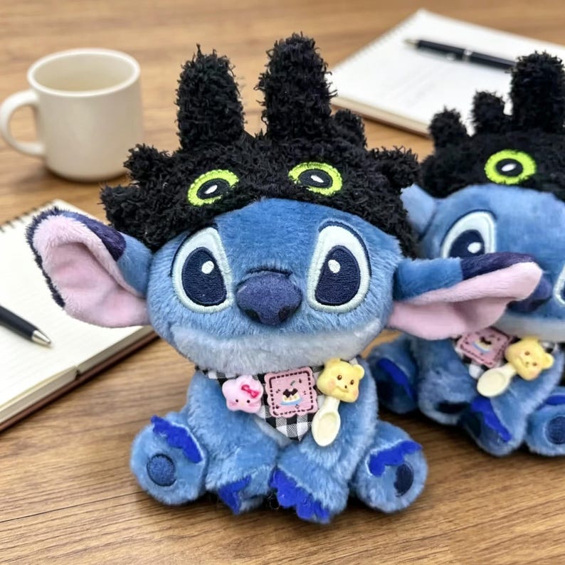 May include: A blue plush character toy wearing a black fuzzy hat with green-rimmed eyes. The toy features large ears, a black and white checkered neck scarf, and small charms including a pink star, a square graphic tag, and a yellow bear spoon.