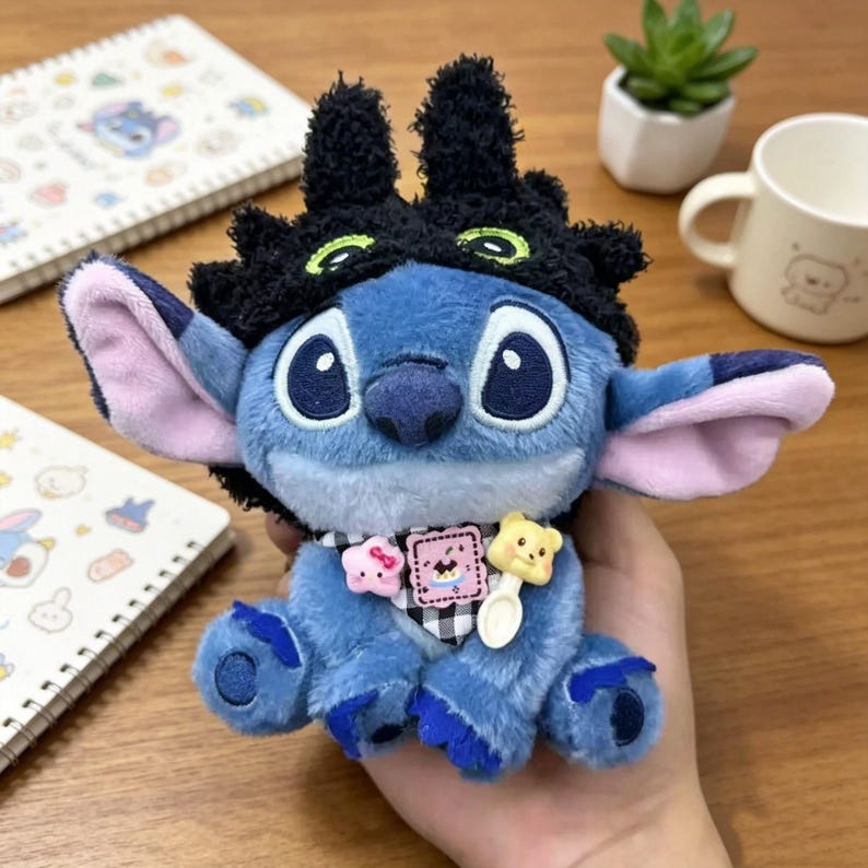 May include: A blue plush toy character with large ears and a black fuzzy headpiece featuring green eye accents. The toy wears a black and white checkered neck accessory adorned with a small pink star, a square graphic patch, and a yellow bear-shaped charm.