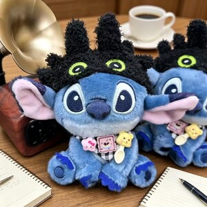 May include: A blue plush character toy wearing a black fuzzy hat with green eyes. The toy features a black and white checkered neck scarf adorned with a small pink star, a square patch with a graphic, and a yellow bear-shaped spoon charm.