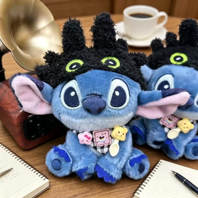 May include: A blue plush character toy wearing a black fuzzy hat with green eyes. The toy features a black and white checkered neck scarf adorned with a small pink star, a square patch with a graphic, and a yellow bear-shaped spoon charm.