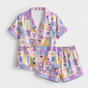 Könnte beinhalten: Ein kurzärmeliges Schlafanzug-Set mit geknöpftem Oberteil und passender Shorts, das ein Patchwork-Muster aus pastellgelben, violetten und blauen Quadraten aufweist. Der Druck enthält Illustrationen von Schmetterlingen, Herzen, Schnecken, Erdbeeren sowie den Text LOVE und XOXO.