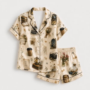 Op de afbeelding: Een crèmekleurige pyjamaset met korte mouwen, knoopsluiting en bijpassende short, voorzien van een herhalende print met vintage illustraties van onder andere stapels boeken, een typemachine, koffiebekers, bloemen, een camera en een schip in een fles.