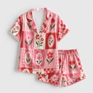 Puede incluir: Un conjunto de pijama de dos piezas que incluye una camisa de manga corta con botones y pantalones cortos a juego. La tela presenta un diseño de patchwork en tonos rosa y rojo, decorado con varias ilustraciones florales, cuadros y líneas onduladas.