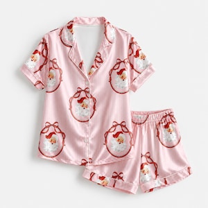 Op de afbeelding: Een pyjamaset met een pyjamashirt met korte mouwen en knoopsluiting en een bijpassende short in lichtroze met dunne witte verticale strepen. De stof heeft een herhalend patroon van vintage kerstmangezichten omlijst door donkerrode strikken.