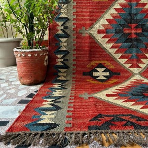 Peut inclure: Un tapis tissé présentant une base rouge vive avec des motifs géométriques en losange dans des tons bleu, crème et jaune moutarde. Le tapis possède une surface texturée, une bordure décorative en zigzag et une frange sur le bord inférieur.