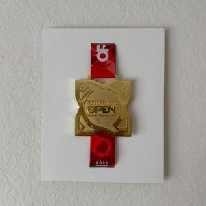May include: A gold-colored medal with the word "OPEN" and the year "2025" on a red ribbon. The medal is mounted on a white square. The ribbon has a logo at the top.