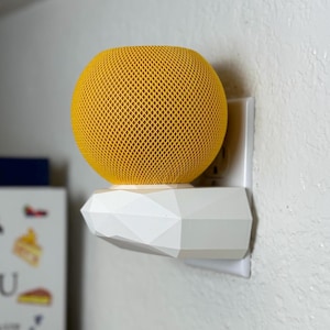 May include: A yellow mesh-covered smart speaker sits atop a white geometric shelf, plugged into a wall outlet. The speaker has a spherical shape. The shelf is a decorative accessory for the speaker, providing a clean, modern look.