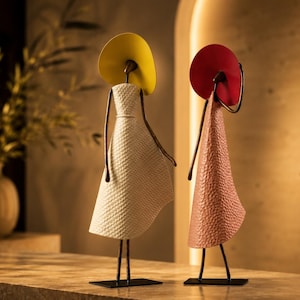 Peut inclure: Deux sculptures abstraites en métal représentant des silhouettes sur des bases rectangulaires. L'une porte un chapeau jaune et une robe blanche texturée, l'autre un chapeau rouge et une robe rose texturée. Les deux ont des membres fins et sombres.