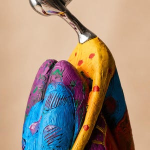 Puede incluir: Una escultura de una figura con cabeza y cuello de plata pulida, inclinada hacia adelante. El cuerpo est&aacute; pintado en amarillo, azul y morado vibrantes con pinceladas texturizadas y lunares rojos, mostrando una postura sentada con rodillas.