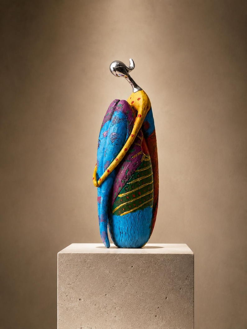 Puede incluir: Una escultura estilizada de una figura con cabeza y cuello de plata pulida, con un cuerpo adornado con patrones vibrantes y multicolores que incluyen azul, amarillo, morado y verde con puntos rojos, representada en una postura sentada y abrazada.
