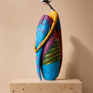Puede incluir: Una escultura abstracta alta de una figura en posici&oacute;n sentada y abrazada. La figura tiene una cabeza y cuello de tono plateado liso, con un cuerpo pintado en patrones vibrantes y multicolores que incluyen azul brillante, amarillo, morado y verde.