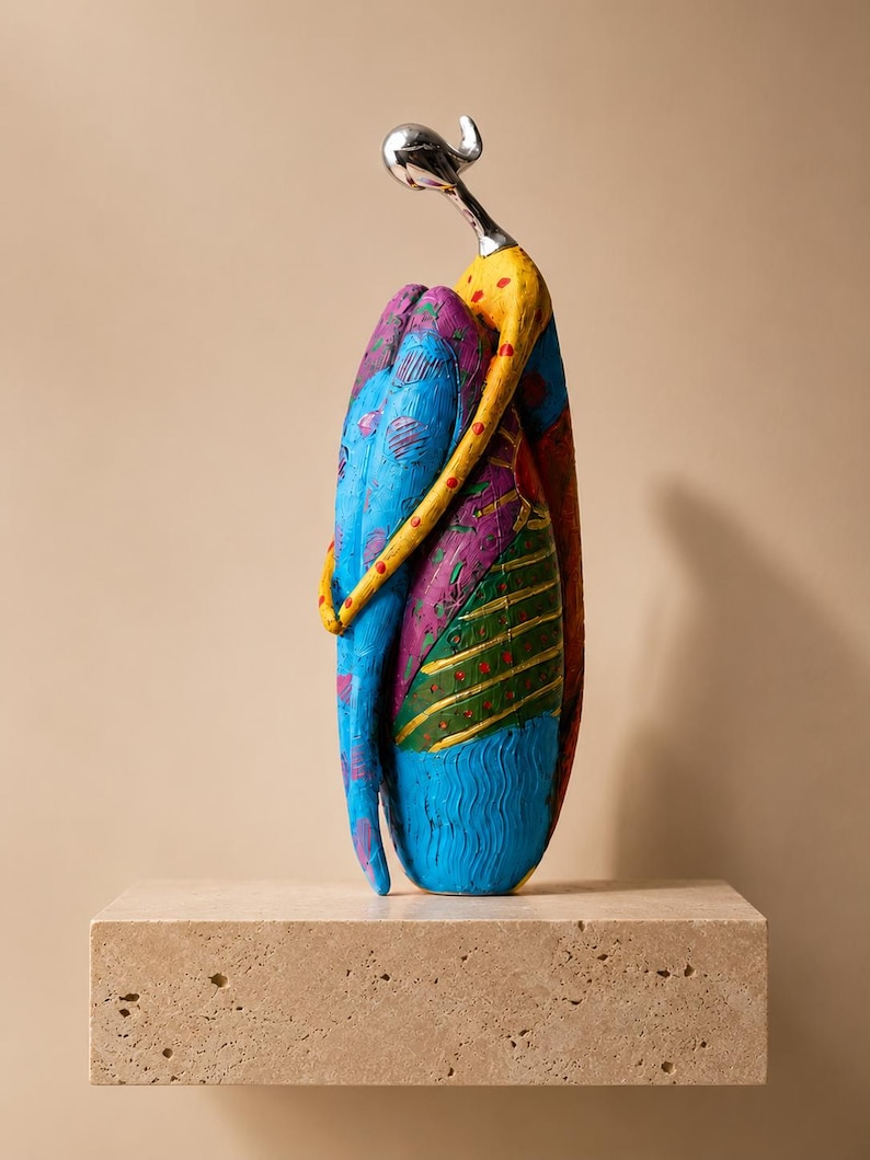 Puede incluir: Una escultura abstracta alta de una figura en posici&oacute;n sentada y abrazada. La figura tiene una cabeza y cuello de tono plateado liso, con un cuerpo pintado en patrones vibrantes y multicolores que incluyen azul brillante, amarillo, morado y verde.