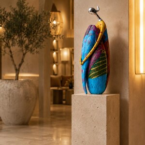 Puede incluir: Una escultura abstracta alta que presenta una figura estilizada con una cabeza lisa de tono plateado y un cuerpo curvo pintado en tonos vibrantes de azul, amarillo, magenta y verde con patrones texturizados y grabados, sobre un pedestal de piedra beige.