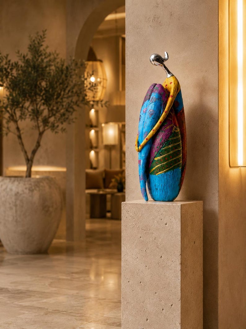 Puede incluir: Una escultura abstracta alta que presenta una figura estilizada con una cabeza lisa de tono plateado y un cuerpo curvo pintado en tonos vibrantes de azul, amarillo, magenta y verde con patrones texturizados y grabados, sobre un pedestal de piedra beige.