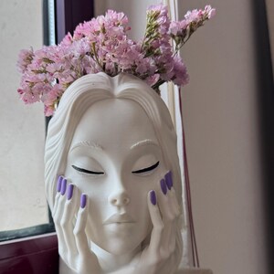 Pode incluir: Um vaso de cerâmica branca em forma de cabeça com um rosto estilizado, olhos fechados e mãos apoiadas nas bochechas. Os dedos têm unhas pintadas de roxo. Um ramo de flores secas cor-de-rosa claro está arranjado na abertura superior.