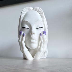 Pode incluir: Uma escultura branca mate de uma cabeça humana com olhos fechados e sobrancelhas definidas. A figura tem as mãos apoiadas em ambas as bochechas, com as unhas pintadas de uma cor violeta vibrante. O cabelo é esculpido com linhas fluidas e visíveis.