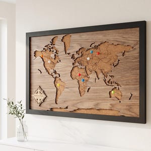 May include: A rectangular world map wall art piece featuring a dark wood frame and a cork backing. The continents are cut from wood and layered over the cork. A compass rose is in the bottom left corner with the text Robinson's Family.