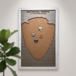 May include: A rectangular wall-mounted cork board with a light grey frame and a central cutout shaped like the National Park Service arrowhead logo. The board features the text NATIONAL PARKS OF THE UNITED STATES and four small circular pins.