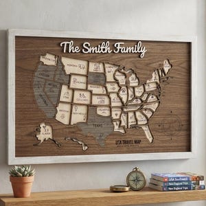 Pode incluir: Uma peça de arte de parede em madeira emoldurada com um mapa em camadas dos Estados Unidos e o texto The Smith Family no topo. Cada estado é cortado e gravado com ícones. O mapa inclui o Alasca e o Havaí, com o texto USA TRAVEL MAP.