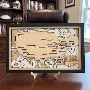 May include: A framed, multi-layered wooden map of the United States featuring state outlines and small football-shaped markers. The background displays white silhouettes of football players and a helmet. A logo reads MapMarkedStudio Stadium Tour.