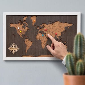 Könnte beinhalten: Ein rechteckiges Weltkarten-Wandbild mit einem dunklen Holzhintergrund und Kork-Kontinenten in einem weißen Rahmen. Die Karte enthält eine Windrose mit dem Text Robinson's Family USA Travel Map und mehrere bunte Pinnnadeln.