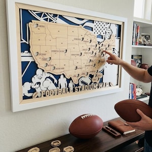 May include: A framed wooden map of the United States featuring cutouts for football stadiums, with the text FOOTBALL STADIUM MAP at the bottom. The map is layered over a blue background with white illustrations of football players and a helmet.