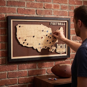 Peut inclure: Une carte en bois encadrée des États-Unis intitulée Football Stadium Map, présentant les contours des États et de petits marqueurs en forme de ballon de football. Un logo en forme de bouclier indique MapMarkedStudio Stadium Tour.