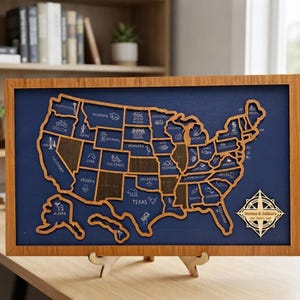 Può includere: Una mappa degli Stati Uniti in una cornice rettangolare di legno, con i contorni degli stati tagliati al laser in legno chiaro su uno sfondo blu scuro. Ogni stato include testo e icone incise. Una rosa dei venti riporta la scritta Serena & Julian's USA Travel Map.