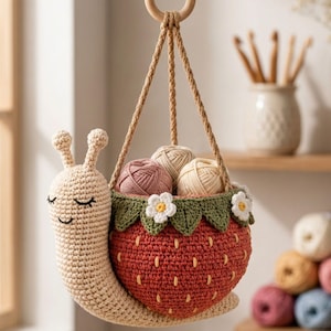 Puede incluir: Un caracol de ganchillo beige con expresión de ojos cerrados, con una concha en forma de fresa roja con hojas verdes y flores blancas. La concha sirve como cesta colgante que contiene tres ovillos de lana, suspendida por un anillo de madera.