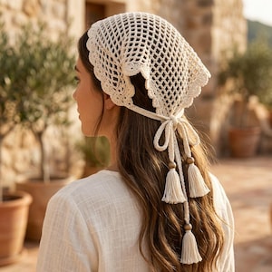 May include: An off-white crochet headscarf featuring a diamond-shaped openwork pattern and a scalloped edge. The headscarf is secured at the back with thin ties adorned with three wooden beads and three matching off-white yarn tassels.