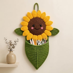 Pode incluir: Um organizador de parede em forma de girassol feito de crochê. O girassol tem pétalas amarelas, um centro castanho com um rosto sorridente e duas folhas verdes. Abaixo, há um bolso verde em forma de folha com várias ferramentas.