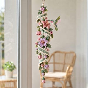 May include: A hanging crochet decoration featuring a spiraling green and white vine adorned with pink and purple flowers, green leaves, and a small hummingbird with green, pink, and white wings. The piece is suspended by a thin string.